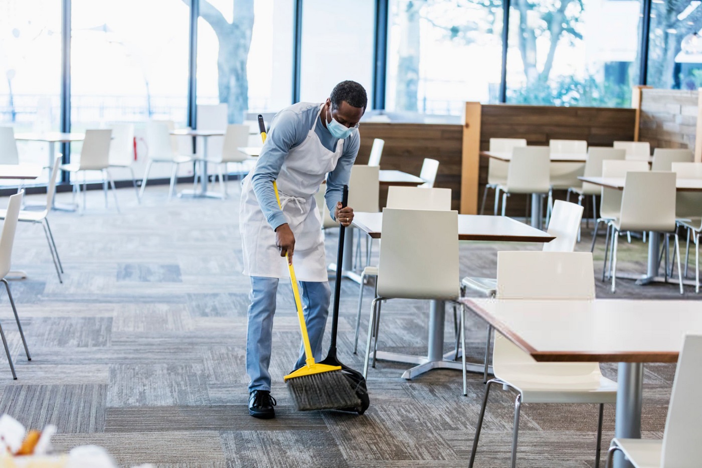 A mid adult African-American man in his 30s working in an empty cafeteria, sweeping the floor. He is wearing a protective face mask, trying to prevent the spread of coronavirus during the COVID-19 pandemic.