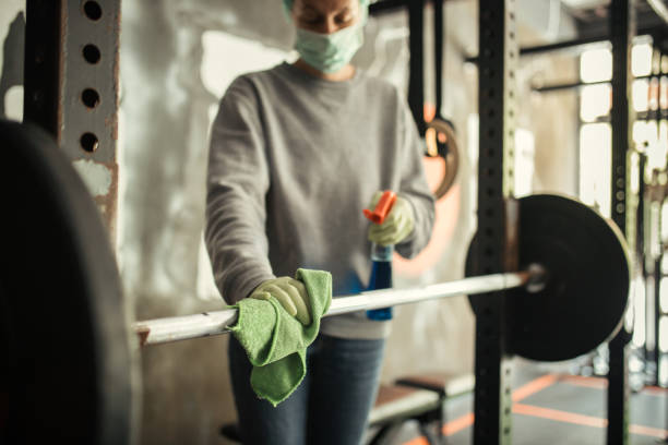 Mature woman cleaning and disinfecting equipment in gym without people. She is using microfiber cloth and antiseptic liquid spray and she wears protective cap, mask and gloves. Disinfection and hygiene in gym is very important to stop spreading virus infections. Photo taken during corona virus outbreak